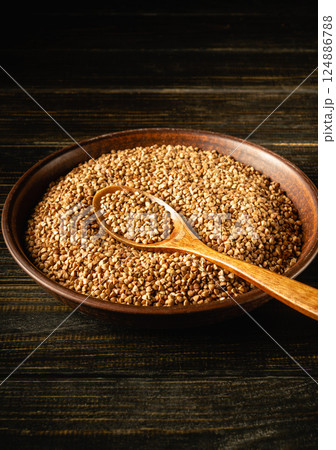 A wooden bowl filled with small brown buckwheat seeds sits on a dark wooden surface. Inside lies a wooden spoon, indicating preparation for cooking or seasoning A wooden bowl filled with small brown buckwheat seeds sits on a dark wooden surface. Inside lies a wooden spoon, indicating preparation for cooking or seasoning 124886788