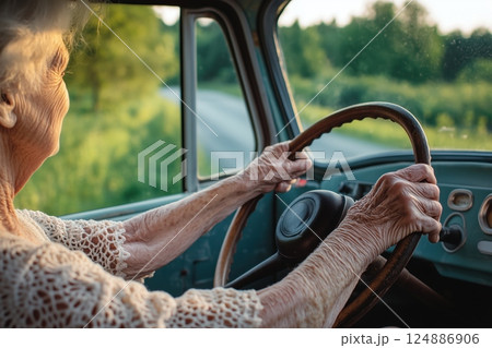 Senior woman drives classic car along sunlit country road POV, wrinkled hands firmly grip large steering wheel, timeless scene shows active senior lifestyle, beautiful senior independence. 124886906