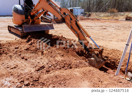 Construction worker uses an excavator to dig trench in dirt field during works construction Construction worker uses an excavator to dig trench in dirt field during works construction 124887518