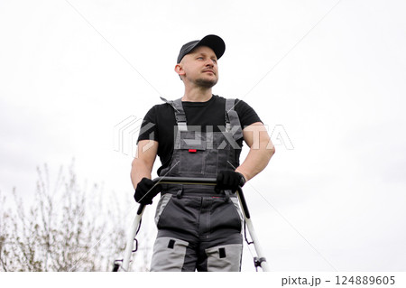 Bottom view of professional male gardener in protective apparel, gloves, cap and overalls holds handle of gasoline cordless lawnmower for mowing grass lawn outdoors. Seasonal landscaping design work Bottom view of professional male gardener in protective apparel, gloves, cap and overalls holds handle of gasoline cordless lawnmower for mowing grass lawn outdoors. Seasonal landscaping design work 124889605