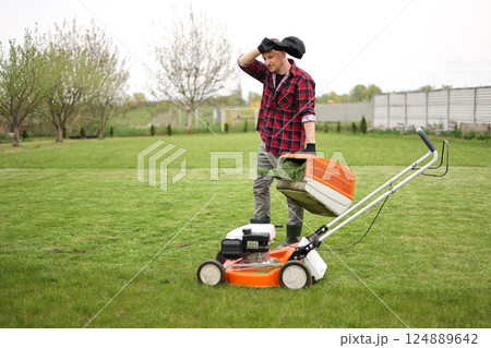 Tired man in casual clothes holds bag with cut grass of lawn mower after mowing lawn with at own backyard. Husband takes care of garden. Modern gasoline garden equipment. Landscaping work at home Tired man in casual clothes holds bag with cut grass of lawn mower after mowing lawn with at own backyard. Husband takes care of garden. Modern gasoline garden equipment. Landscaping work at home 124889642