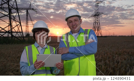 Man and woman engineers with tablet smile to camera against electric power generation plant in sunset field. Engineers work together at power distribution substation at twilight time closeup Man and woman engineers with tablet smile to camera against electric power generation plant in sunset field. Engineers work together at power distribution substation at twilight time closeup 124890431