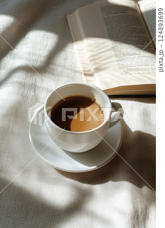 Warm coffee cup beside an open book on a sunlit table with leafy shadows 124893699