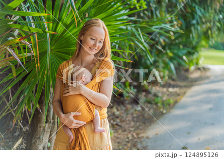 Mother holding her little baby in a yellow sling in the park. Warm and loving family moment. Babywearing, parenting, and mother-child bonding concept Mother holding her little baby in a yellow sling in the park. Warm and loving family moment. Babywearing, parenting, and mother-child bonding concept 124895126