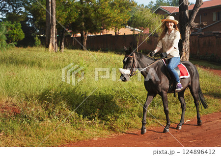 Young woman in shirt and straw hat, riding black horse in the park, blurred background with houses and trees Young woman in shirt and straw hat, riding black horse in the park, blurred background with houses and trees 124896412