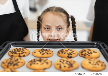 Caucasian young girl with braided hair, wearing apron, smiling, displaying baked chocolate chip cookies, surrounded by family members engaged in baking activity. 124902806