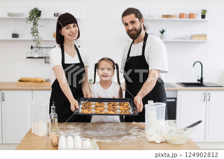 Caucasian smiling family consisting of parents and young daughter presenting baked cookies in kitchen. Scene conveys family bonding, baking activity, happiness, and warm atmosphere. Caucasian smiling family consisting of parents and young daughter presenting baked cookies in kitchen. Scene conveys family bonding, baking activity, happiness, and warm atmosphere. 124902812