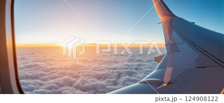 Airplane window view of sunset above fluffy clouds, with aircraft wing in foreground. The warm glow of the setting sun creates a serene and peaceful atmosphere 124908122