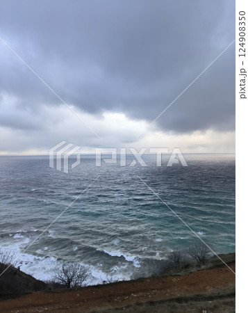 Dramatic sky with large grey clouds above choppy ocean waves, viewed from a grassy cliff edge. The sea has a gradient of colors from deep blue to lighter teal near the shore 124908350