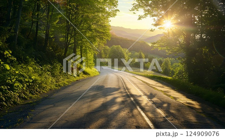 The setting sun lights up a green forest and a winding asphalt road in a wide panoramic view 124909076