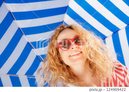 Happy young woman under a striped blue parasol. Girl wearing sunglasses enjoys a summer vacation 124909422