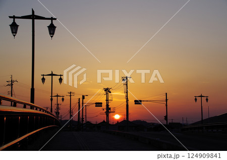 レトロな街灯が設置された橋の歩道から遥か彼方の空に浮かんだ夕日を眺める春の穏やかな夕暮れの場面 124909841
