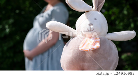Soft plush bunny toy in focus with a blurred pregnant woman holding her belly in the background 124910985