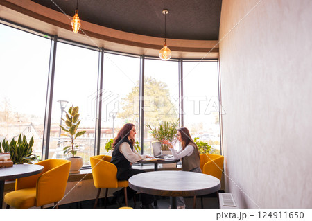 Two smartly dressed young Caucasian women sitting at a cafe and having a deep conversation about marketing strategies. Concept of online business growth and professional cooperation. Wide shot 124911650