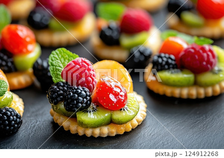 Colorful fruit tarts arranged on a dark slate surface. Each tart consists of a small round biscuit base topped with an assortment of fresh fruits including raspberries, blackberries, kiwi slices. 124912268