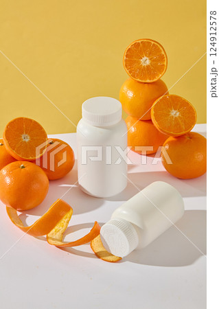 The white medicine bottle stands on the table while another bottle lies next to the wavy orange peel. Citrus are stacked on top of each other, presented on a white plate, light brown background. 124912578