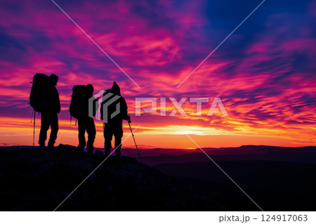Three people are standing on a mountain top, looking out at the sunset 124917063