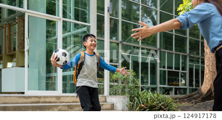 Joy and Embrace. A young boy runs towards his mother after school, holding a soccer ball, expressing pure happiness. 124917178