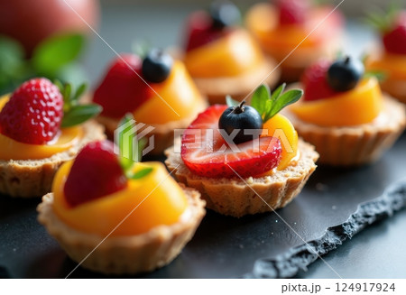 Close-up view of beautifully arranged mini fruit tarts on a dark surface. Each tart features a golden pastry shell filled with cream and topped with colorful fresh fruits including strawberries. 124917924