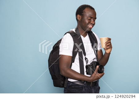 African American man touches his camera lens, smiling as he looks at the disposable cup in his hand. Black male photographer with a backpack and DSLR gazes at his coffee with a content expression. 124920294