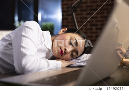 Closeup of asian businesswoman sleeping at desk during her night shift, with her laptop and office workspace still active. Her exhaustion is visible as she rests on her desk after hours of work. 124920334