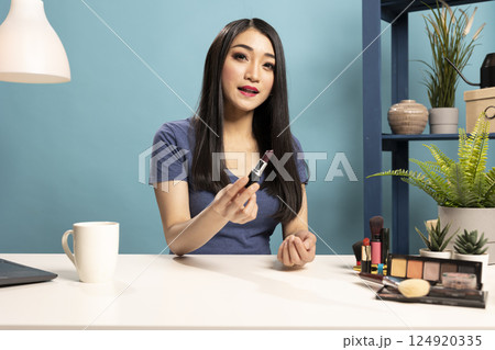 Asian woman recording a beauty tutorial video at desk, presenting lipstick shade to her camera. Beauty influencer holds a cosmetic product as she speaks to online viewers in live makeup demonstration. Asian woman recording a beauty tutorial video at desk, presenting lipstick shade to her camera. Beauty influencer holds a cosmetic product as she speaks to online viewers in live makeup demonstration. 124920335