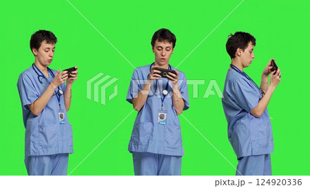 Medical assistant playing mobile video games on smartphone app, taking a break from healthcare work. Nurse enjoys gaming tournament on phone, standing against greenscreen backdrop. Camera B. Medical assistant playing mobile video games on smartphone app, taking a break from healthcare work. Nurse enjoys gaming tournament on phone, standing against greenscreen backdrop. Camera B. 124920336