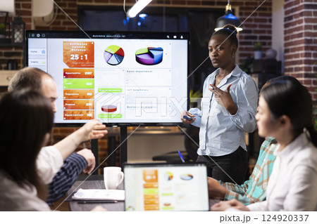 Professional black woman stands near digital monitor, presenting business analytics updates with her colleagues. Multiethnic startup team collaborating in brick wall office, working late at night. 124920337