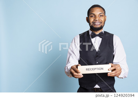 Hotel concierge presenting the reception indicator mark at front desk, welcoming clients at five star holiday resort in studio. Professional doorkeeper holds wall sign against blue background. Hotel concierge presenting the reception indicator mark at front desk, welcoming clients at five star holiday resort in studio. Professional doorkeeper holds wall sign against blue background. 124920394