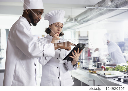 Team of diverse experts researching new cooking tips and recipes on tablet, preparing gourmet meals in the restaurant kitchen. Professional chefs read about combinations of ingredients. 124920395