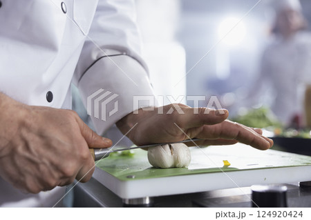 Sous chef pressing knife on cloves to make crushed garlic on the cutting board, creating condiments with a fresh flavor. Young cook preparing a gourmet dish at the counter, chef life. Close up. Sous chef pressing knife on cloves to make crushed garlic on the cutting board, creating condiments with a fresh flavor. Young cook preparing a gourmet dish at the counter, chef life. Close up. 124920424