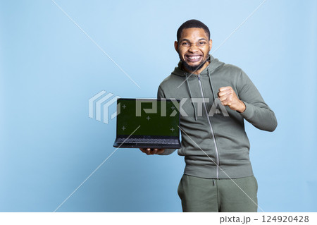 Confident happy person pointing at green screen mockup in studio, using personal laptop for an advertisement on camera. African american young guy gesturing against blue background. Confident happy person pointing at green screen mockup in studio, using personal laptop for an advertisement on camera. African american young guy gesturing against blue background. 124920428