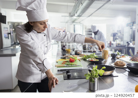 In a modern kitchen, woman head chef adding herbs to complete a gourmet dish, balancing flavors with rosemary and other greenery. Kitchen counter stacked with vegetables and food prep. 124920457