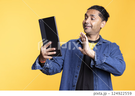 Radiant young adult saluting his friend over online video call connection, showcases a friendly reaction. Joyful guy communicating with a person on teleconference using tablet. Radiant young adult saluting his friend over online video call connection, showcases a friendly reaction. Joyful guy communicating with a person on teleconference using tablet. 124922559