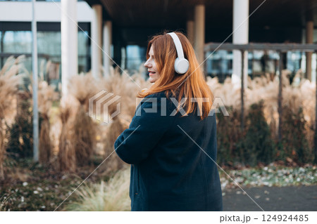 Beautiful young happy woman in headphones with bouquet of tulip posing on urban background. Smiling 30s girl listening to music through wireless headphones, rear view Beautiful young happy woman in headphones with bouquet of tulip posing on urban background. Smiling 30s girl listening to music through wireless headphones, rear view 124924485