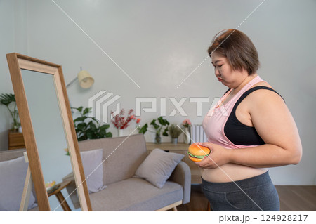 Pensive woman holding a burger, gazing at her reflection in a mirror, contemplating her body image and choices 124928217