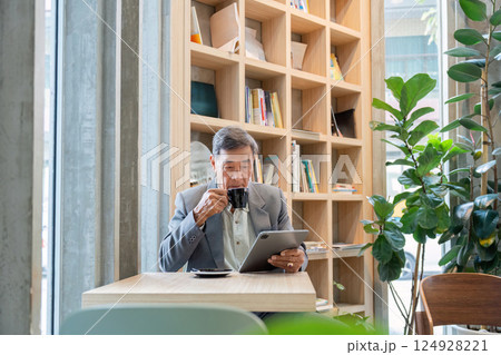 Elderly man sipping coffee while reading a tablet in a vibrant cafe filled with greenery 124928221