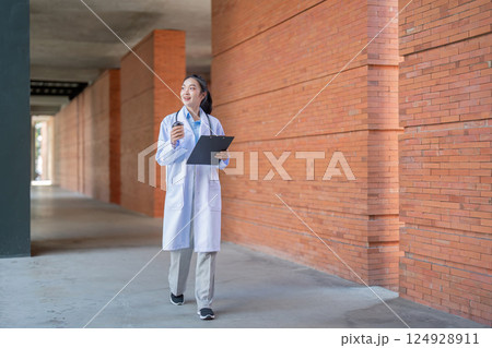 Asian female doctor walking with coffee and clipboard in hospital corridor Asian female doctor walking with coffee and clipboard in hospital corridor 124928911