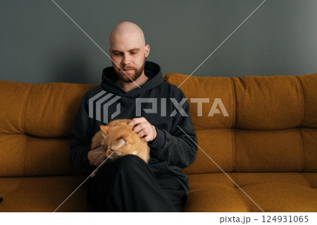 Portrait of young bald man with beard wearing casual clothes sitting on comfortable sofa, lovingly petting ginger cat, enjoying quiet moment at home. Concept of relationship between people and animal. 124931065