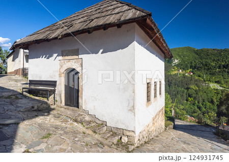 Jajce town in Bosnia and Herzegovina, Europe. Quaint white building with a dark wooden door and traditional roof, nestled in a hillside village under a bright blue sky. Jajce town in Bosnia and Herzegovina, Europe. Quaint white building with a dark wooden door and traditional roof, nestled in a hillside village under a bright blue sky. 124931745