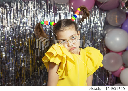 Offended girl in glasses and yellow holiday blouse with funny ponytails hairstyle on festive background with balloons 124931840