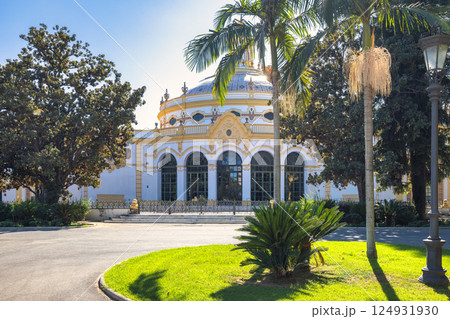 Seville town, capital of Andalusia in Spain. Elegant rotunda building in a sunny park setting, surrounded by lush greenery and palm trees. 124931930