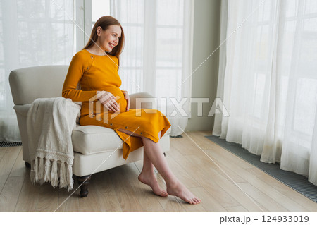 Pregnancy motherhood people expectation future. Pregnant woman with big belly sitting on chair near window at home. Girl hugging her tummy enjoying pregnancy. Maternity tenderness parenthood new life 124933019