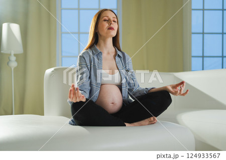 Pregnancy prenatal breathing. Pregnant woman with big belly sitting in lotus pose on sofa at home. Pregnant girl practicing yoga mindfulness meditation. Relaxation self care during pregnancy 124933567