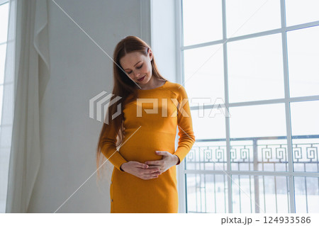 Pregnancy motherhood people expectation future. Pregnant woman with big belly standing near window at home. Girl hugging her tummy enjoying pregnancy. Maternity tenderness parenthood new life concept 124933586