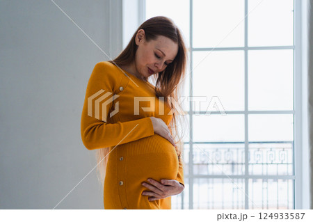 Pregnancy motherhood people expectation future. Pregnant woman with big belly standing near window at home. Girl hugging her tummy enjoying pregnancy. Maternity tenderness parenthood new life concept 124933587