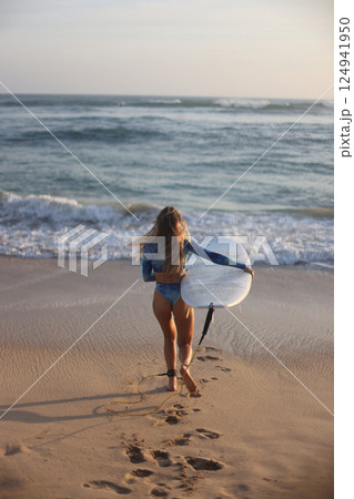 A woman is leisurely walking along the beach while holding a surfboard 124941950