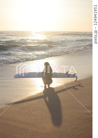 A Surfer Woman Can Be Seen Strolling Along the Beach as the Sun Sets in the Beautiful Sky 124941951