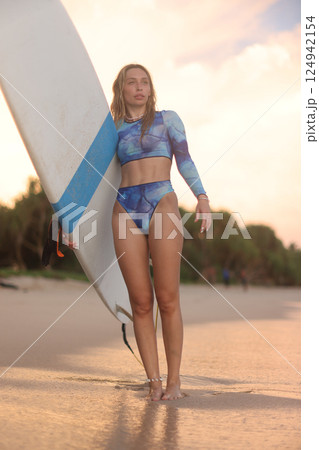 A woman wearing a bikini is standing on the beach while holding a surfboard A woman wearing a bikini is standing on the beach while holding a surfboard 124942154