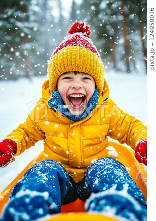 A young boy in a yellow jacket and red hat is sledding down a hill in the snow A young boy in a yellow jacket and red hat is sledding down a hill in the snow 124950016